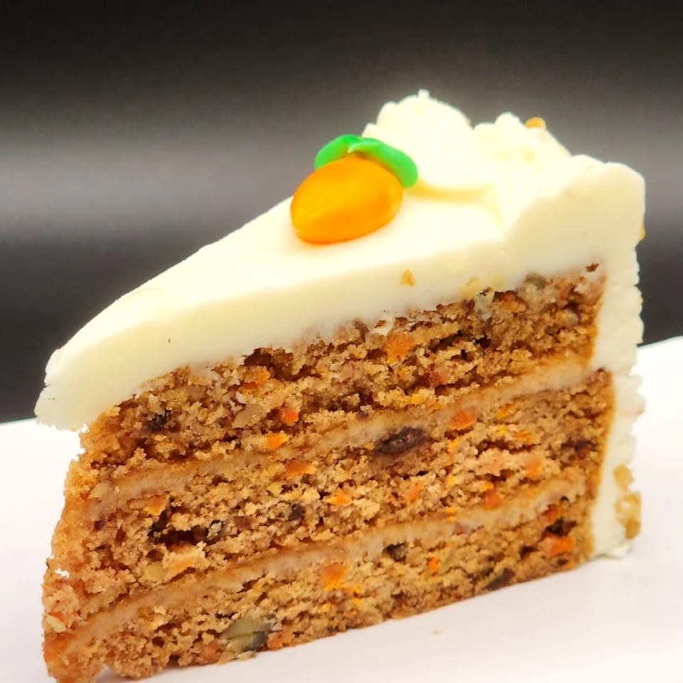 A slice of carrot cake with white cream cheese frosting and a small orange carrot decoration.