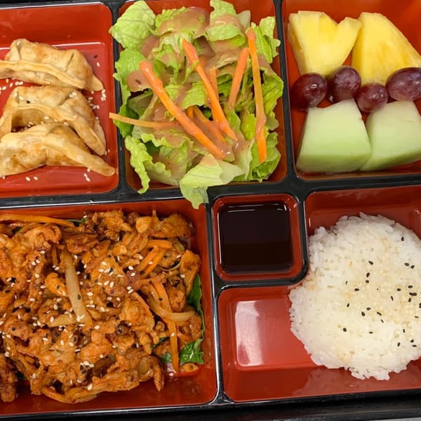 A bento box with dumplings, salad, fruit, spicy chicken, rice, and dipping sauce.