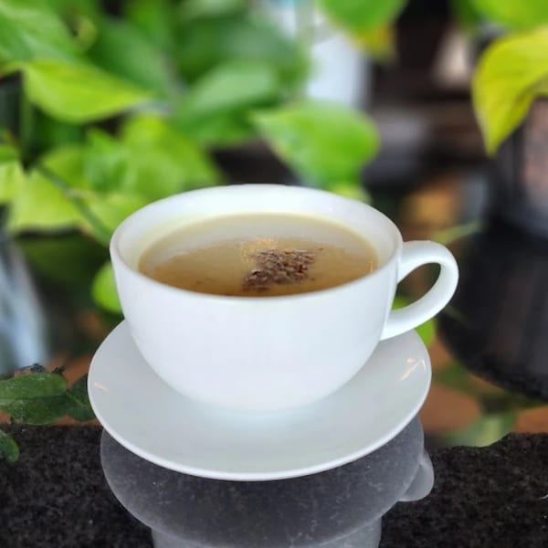 A white cup of coffee with a sprinkle of spices on top, set against a green leafy background.